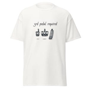 May include: White t-shirt with the text "3rd pedal required" above an illustration of three car pedals. The pedals are in shades of gray and black, with a textured appearance. The shirt is a casual, unisex style.