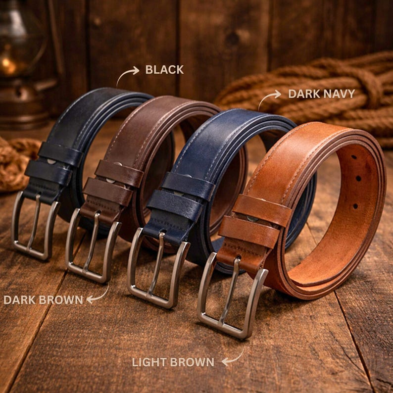 May include: Four leather belts in black, dark brown, dark navy, and light brown, each with a silver-colored buckle. The belts are arranged on a wooden surface, with text labels indicating the color of each belt.