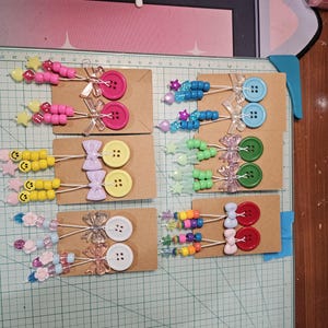 May include: A collection of handmade beaded hair accessories, including colorful beaded sticks with buttons, bows, and star-shaped beads. The accessories are arranged on small cardboard cards, showcasing a variety of colors and designs.