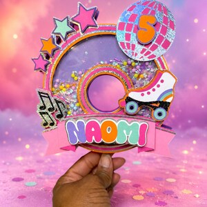 May include: A circular shaker cake topper featuring the name NAOMI in colorful letters, a roller skate, musical notes, and stars. A silver disco ball with the number 5 sits at the top. The center is filled with small beads and sequins.