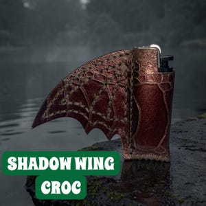 Peut inclure: Un étui de briquet en cuir marron avec un motif en forme d'aile et le texte "SHADOW WING CROC" dans un encadré vert. L'aile présente un motif cousu et un aspect texturé. Le briquet est posé sur un fond naturel flou.