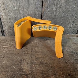 May include: Bright orange 3D-printed ammunition holder with a hinged lid. The holder is designed to hold multiple rounds of ammunition. The text "358 WIN" is printed on the side of the holder. The holder is sitting on a wooden surface.