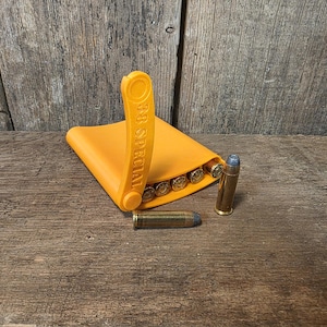 May include: An orange 38 Special speed loader with several bullets inside. Two additional bullets are next to the speed loader. The speed loader and bullets are on a wooden surface.