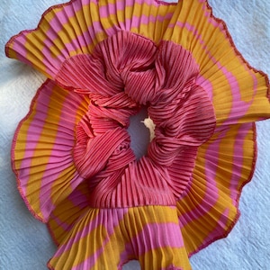 May include: A circular hair scrunchie featuring a pleated fabric design with alternating horizontal stripes of bright pink and golden yellow. The outer edge of the ruffled fabric is finished with a thin, decorative pink stitched trim.