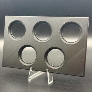 May include: A black rectangular palette with six circular wells, likely for makeup or art supplies. The palette is made of a matte material and is displayed on a clear acrylic stand, showcasing its design and functionality.