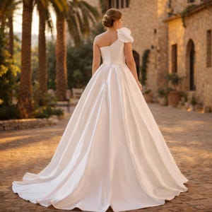 Peut inclure: Robe de mari&eacute;e blanche &agrave; une &eacute;paule, orn&eacute;e d'une grande fleur sur l'&eacute;paule. La robe pr&eacute;sente un corsage ajust&eacute; et une jupe fluide qui tra&icirc;ne derri&egrave;re. L'image est prise &agrave; l'ext&eacute;rieur, avec des palmiers et un b&acirc;timent en pierre en arri&egrave;re-plan.