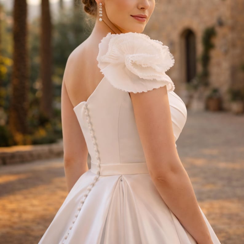Peut inclure: Robe de mari&eacute;e blanche &agrave; une &eacute;paule, orn&eacute;e d'un grand motif floral &agrave; volants sur l'&eacute;paule. La robe pr&eacute;sente un corsage ajust&eacute; avec une rang&eacute;e de boutons dans le dos et une jupe fluide. Le fond est flou.