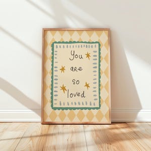以下が含まれることがあります： 「You are so loved」の文字が黒い筆記体で書かれ、金色の星がアクセントになった額入りのプリント。クリーム色の背景に緑色の縁取りがあり、黄色のひし形模様の上に配置されています。