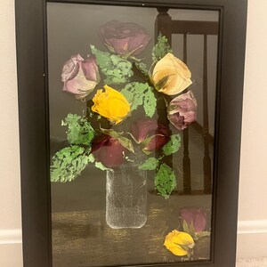 May include: A framed artwork featuring a bouquet of dried roses in a clear glass vase. The roses are various colors, including yellow, purple, and burgundy, with green leaves. The artwork is set against a dark background and framed in black.