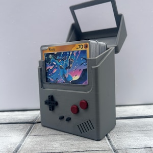 May include: A grey 3D-printed card holder, designed to resemble a retro handheld games console. The holder is open, displaying a stack of trading cards, with one card showing a blue and yellow character. Red buttons and a directional pad are visible.