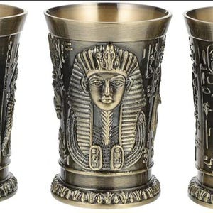 Könnte beinhalten: Drei Metallbecher mit antikem Messing-Finish, jeweils mit geprägten ägyptischen Motiven. Der linke Becher zeigt musizierende Figuren, der mittlere eine Pharaonenmaske und der rechte eine Gottheit mit Schakalkopf.