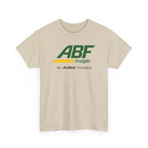 以下が含まれることがあります： ベージュ色のTシャツ。ABF Freightのロゴが緑と黄色でプリントされています。「Freight」の文字がABFロゴの下にあり、「An ArcBest Company」がその下に印刷されています。Tシャツは半袖で、クルーネックです。