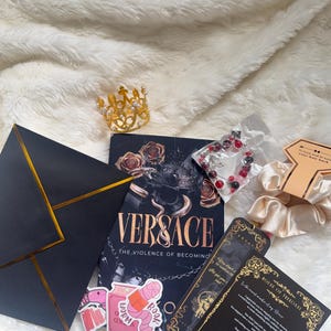 May include: An assortment of items including a book titled "VERSACE: The Violence of Becoming", a gold crown, stickers, a bracelet, and a black envelope with gold trim. The items are arranged on a white, fluffy surface.