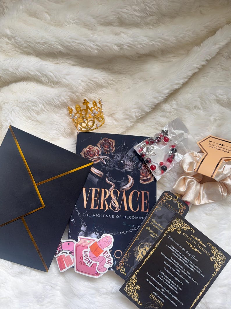 May include: An assortment of items including a book titled "VERSACE: The Violence of Becoming", a gold crown, stickers, a bracelet, and a black envelope with gold trim. The items are arranged on a white, fluffy surface.