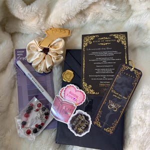 May include: A collection of items including a scrunchie, stickers, a bracelet, a bookmark, and a highlighter. The scrunchie is gold, and the stickers say "Book Lover" and "Dark Romance Reader". The bookmark has a butterfly design.