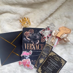 May include: An assortment of items including a book titled "Versace", a gold crown, stickers, a bookmark, a scrunchie, a pen, a black envelope with gold trim, and a beaded bracelet. The items are arranged on a white, fluffy surface.