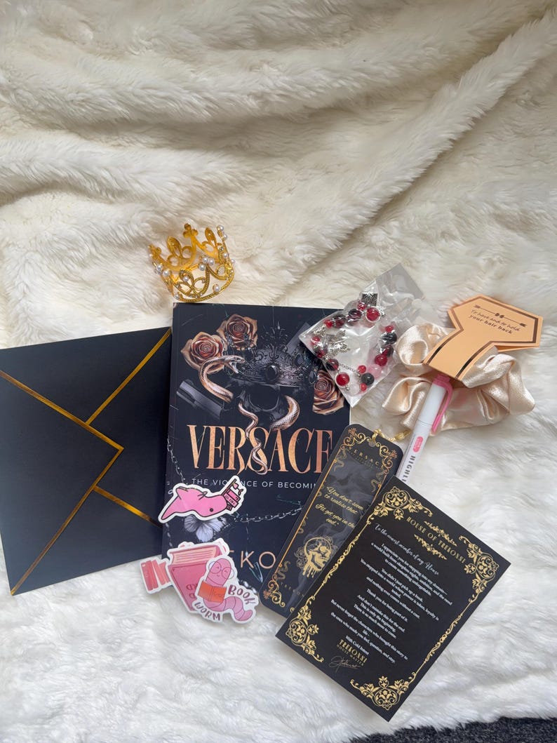 May include: An assortment of items including a book titled "Versace", a gold crown, stickers, a bookmark, a scrunchie, a pen, a black envelope with gold trim, and a beaded bracelet. The items are arranged on a white, fluffy surface.