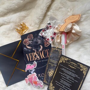 May include: An assortment of items including a book titled "Versace", a gold crown, a scrunchie, a pen, stickers, a black envelope with gold trim, a bookmark, and a card with gold accents, all on a white fluffy surface.