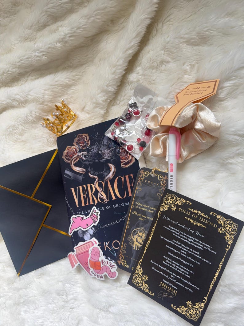 May include: An assortment of items including a book titled "Versace", a gold crown, a scrunchie, a pen, stickers, a black envelope with gold trim, a bookmark, and a card with gold accents, all on a white fluffy surface.