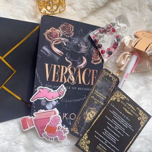 May include: A collection of items including a book titled "VERSACE: The Violence of Becoming", a gold crown, stickers, a bracelet, and a bookmark. The book has a black cover with gold lettering and a snake and rose design. The stickers feature a pink worm reading a book.