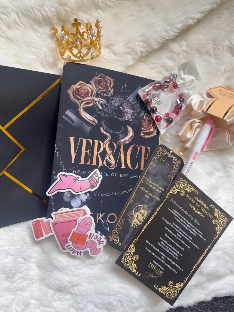 May include: A collection of items including a book titled "VERSACE: The Violence of Becoming", a gold crown, stickers, a bracelet, and a bookmark. The book has a black cover with gold lettering and a snake and rose design. The stickers feature a pink worm reading a book.