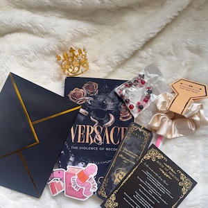 May include: A collection of items including a gold crown, a book titled "VERSACE: The Violence of Becoming", stickers, a bracelet, a scrunchie, and cards with gold accents. The items are arranged on a white, fluffy surface.