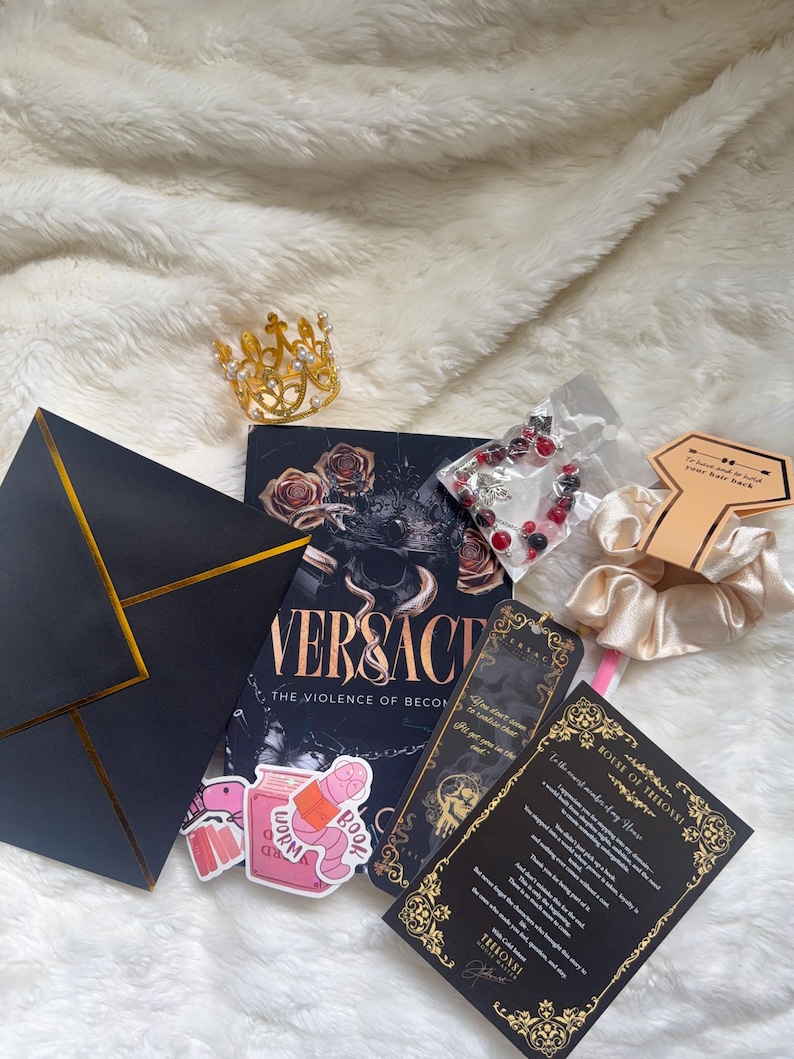 May include: A collection of items including a gold crown, a book titled "VERSACE: The Violence of Becoming", stickers, a bracelet, a scrunchie, and cards with gold accents. The items are arranged on a white, fluffy surface.