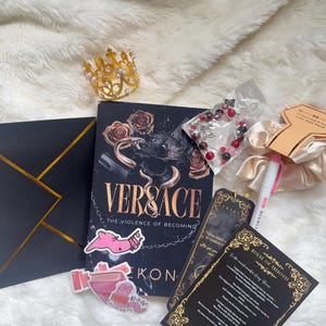 May include: A collection of items including a book titled "Versace: The Violence of Becoming", a gold crown, stickers, a bracelet, and cards with gold accents, all arranged on a fluffy white surface.