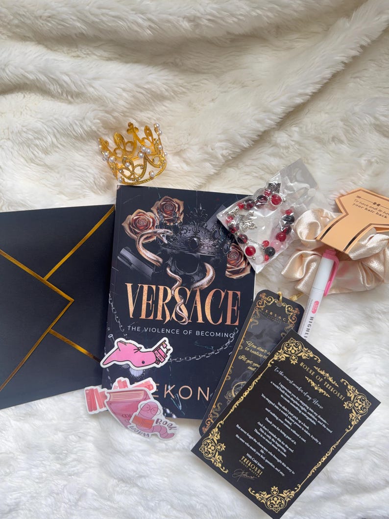 May include: A collection of items including a book titled "Versace: The Violence of Becoming", a gold crown, stickers, a bracelet, and cards with gold accents, all arranged on a fluffy white surface.