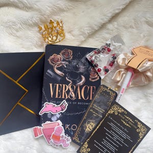 May include: An assortment of items including a book titled "Versace", a gold crown, stickers, a bracelet, and a bookmark. The book has a black cover with gold lettering and rose illustrations. The stickers feature a pink worm and a book. The items are arranged on a white, fluffy surface.