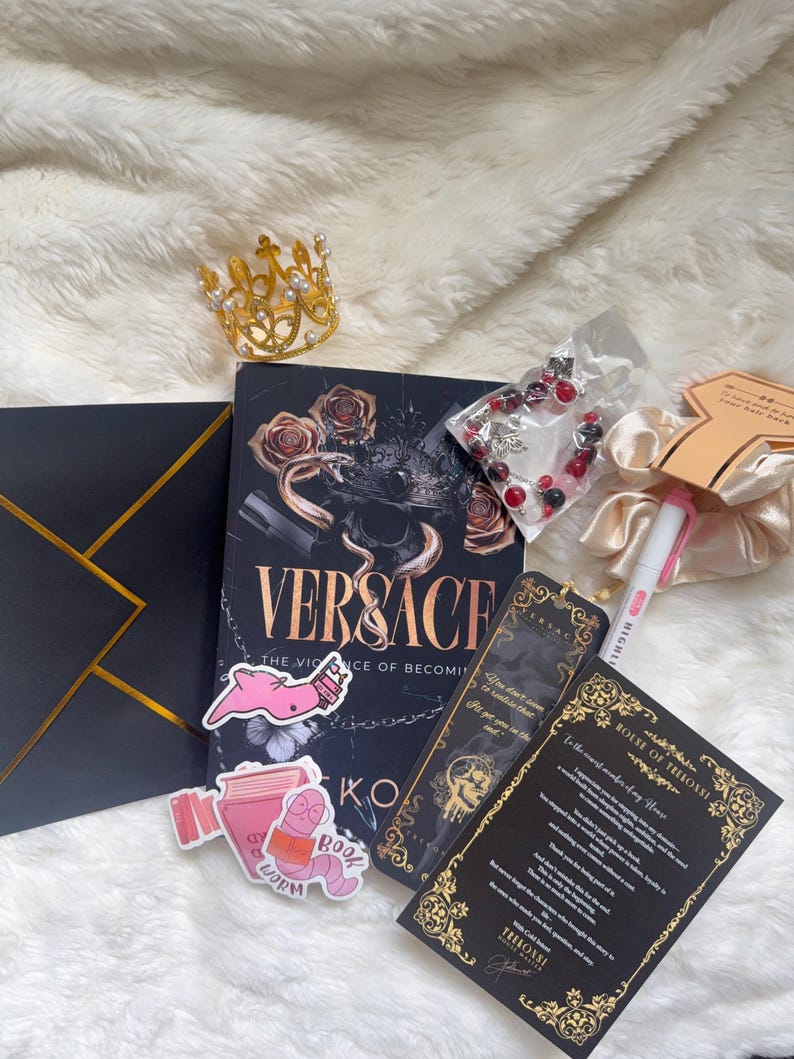 May include: An assortment of items including a book titled "Versace", a gold crown, stickers, a bracelet, and a bookmark. The book has a black cover with gold lettering and rose illustrations. The stickers feature a pink worm and a book. The items are arranged on a white, fluffy surface.