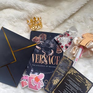 May include: A collection of items including a book titled "VERSACE: The Violence of Becoming", a gold crown, a bracelet, stickers, bookmarks, a pen, and a scrunchie. The items are arranged on a white, fluffy surface.