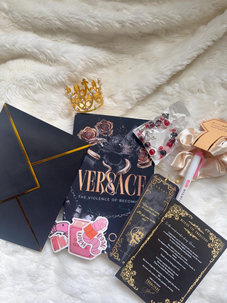 May include: A collection of items including a book titled "VERSACE: The Violence of Becoming", a gold crown, a bracelet, stickers, bookmarks, a pen, and a scrunchie. The items are arranged on a white, fluffy surface.
