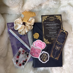 May include: A collection of items including a gold-colored diamond-shaped charm, a cream-colored scrunchie, a purple sticky note pad, a highlighter, a bracelet with red and black beads, and stickers that say "Book Lover" and "Dark Reader".