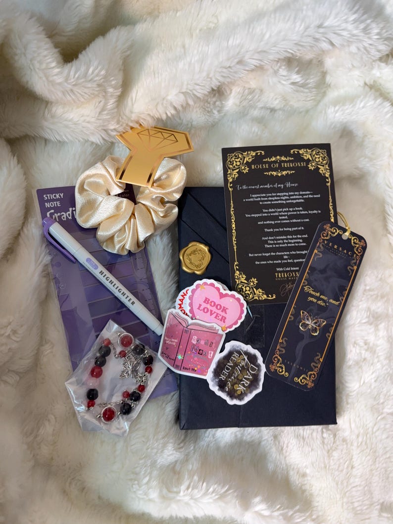 May include: A collection of items including a gold-colored diamond-shaped charm, a cream-colored scrunchie, a purple sticky note pad, a highlighter, a bracelet with red and black beads, and stickers that say "Book Lover" and "Dark Reader".