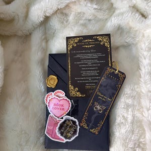 May include: A dark navy gift box with a gold wax seal, stickers, a bookmark, and a card. The stickers read "Romance Addict", "Book Lover", and "Dark Reader". The bookmark has a butterfly design and the text "Touch me, and you die".
