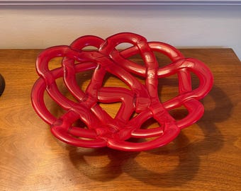 May include: A decorative red glass bowl with a woven, intertwined design. The bowl is oval-shaped and sits on a wooden surface. The bowl's glossy finish reflects light, highlighting its intricate form.
