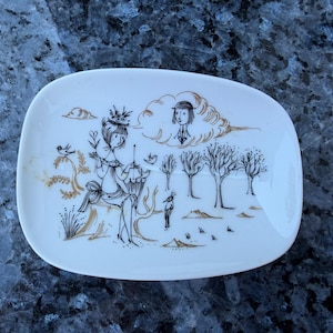 May include: A white ceramic dish featuring a black and gold line drawing of a person sitting on a tree stump holding an umbrella, with a bird, trees, and a person's face inside a cloud depicted in the background. The dish has a rounded rectangular shape.