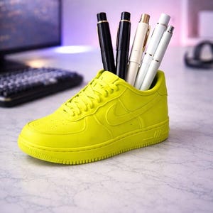 Puede incluir: Un portalápices amarillo neón con forma de zapatilla deportiva, que contiene varios bolígrafos. Los bolígrafos son negros, blancos y dorados. La zapatilla tiene un diseño clásico con el logotipo de la marca en el lateral. El portalápices está sobre una superficie de mármol.