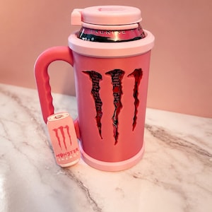 K&ouml;nnte beinhalten: Ein hellrosa Dosenhalter mit Griff und passendem Deckel. Der Halter verf&uuml;gt &uuml;ber ein ausgeschnittenes Design in Form des Monster Energy Logos, das eine rosa-schwarze Getr&auml;nkedose im Inneren zeigt. Ein kleiner rosa Anh&auml;nger in Dosenform ist befestigt.