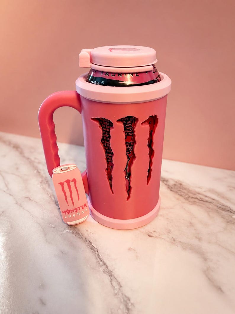 K&ouml;nnte beinhalten: Ein hellrosa Dosenhalter mit Griff und passendem Deckel. Der Halter verf&uuml;gt &uuml;ber ein ausgeschnittenes Design in Form des Monster Energy Logos, das eine rosa-schwarze Getr&auml;nkedose im Inneren zeigt. Ein kleiner rosa Anh&auml;nger in Dosenform ist befestigt.