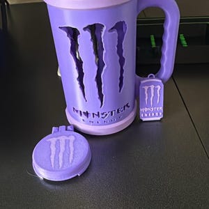 K&ouml;nnte beinhalten: Ein hellvioletter Becher mit dem ausgeschnittenen Monster Energy-Logo auf der Vorderseite und dem darunter aufgedruckten Text Monster Energy. Ein kleiner passender violetter Anh&auml;nger mit dem Logo h&auml;ngt am Griff, ein runder Deckel liegt daneben.