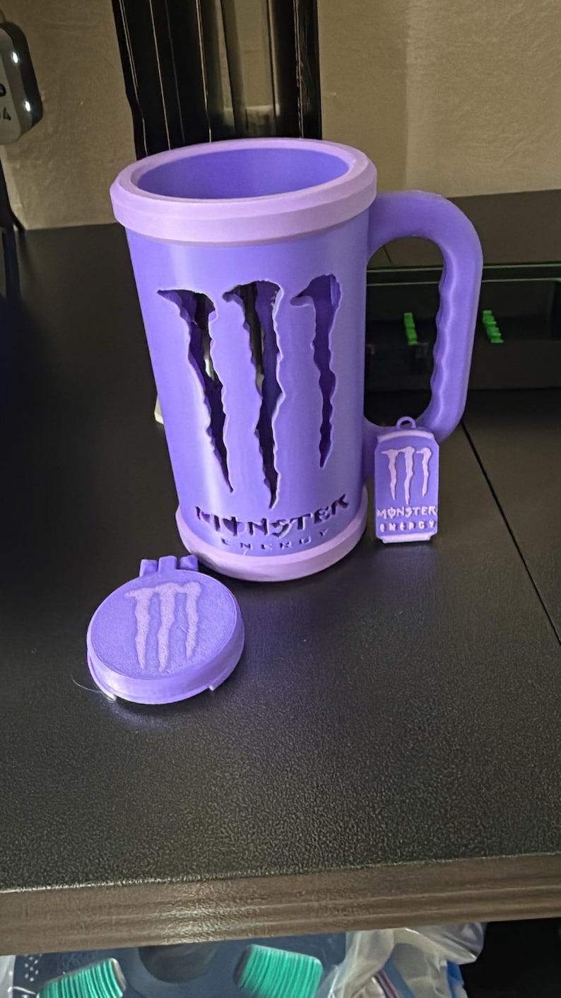 K&ouml;nnte beinhalten: Ein hellvioletter Becher mit dem ausgeschnittenen Monster Energy-Logo auf der Vorderseite und dem darunter aufgedruckten Text Monster Energy. Ein kleiner passender violetter Anh&auml;nger mit dem Logo h&auml;ngt am Griff, ein runder Deckel liegt daneben.