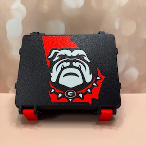 May include: A black rectangular storage case featuring a red silhouette of the state of Georgia on the front. Centered on the red shape is a white and black bulldog head graphic with a spiked collar and a red letter G logo on the collar.