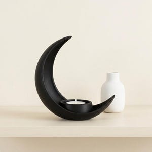 Puede incluir: Un portavelas negro en forma de media luna con una vela pequeña y redonda. A la derecha hay un jarrón blanco minimalista. El portavelas está hecho de un material mate y se encuentra sobre una superficie clara.