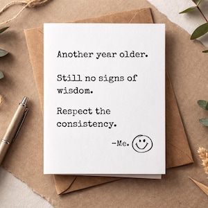 Może przedstawiać: Biała kartka z życzeniami z tekstem "Another year older. Still no signs of wisdom. Respect the consistency." i emotikonem uśmiechu. Karta leży na brązowej kopercie, z długopisem i elementami dekoracyjnymi.