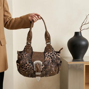 Puede incluir: Un bolso de mano marrón con estampado de leopardo, acentuado con adornos de piel sintética marrón oscuro y tachuelas metálicas plateadas. El bolso incluye un llamativo cierre de hebilla decorativa plateada en la parte frontal y dos asas resistentes.