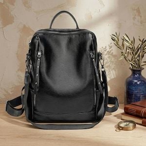 Puede incluir: Una mochila negra con superficie texturizada, que cuenta con un asa superior, correas ajustables para los hombros y dos bolsillos frontales verticales con cremallera y tiradores largos con tachuelas. El compartimento principal tiene cierre de cremallera.