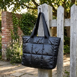 May include: A black quilted tote bag with sturdy black handles hangs from a wooden post. The bag has a rectangular shape with a quilted pattern. The background features a wooden fence, brick wall, and greenery.