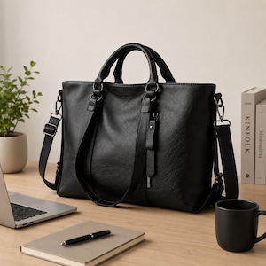 May include: Black leather tote bag with handles and a detachable shoulder strap. The bag has a textured surface and metal hardware. A laptop, notebook, pen, and coffee mug are on the wooden surface.